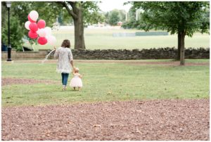 baby girl and mom walking on grass with pink balloons