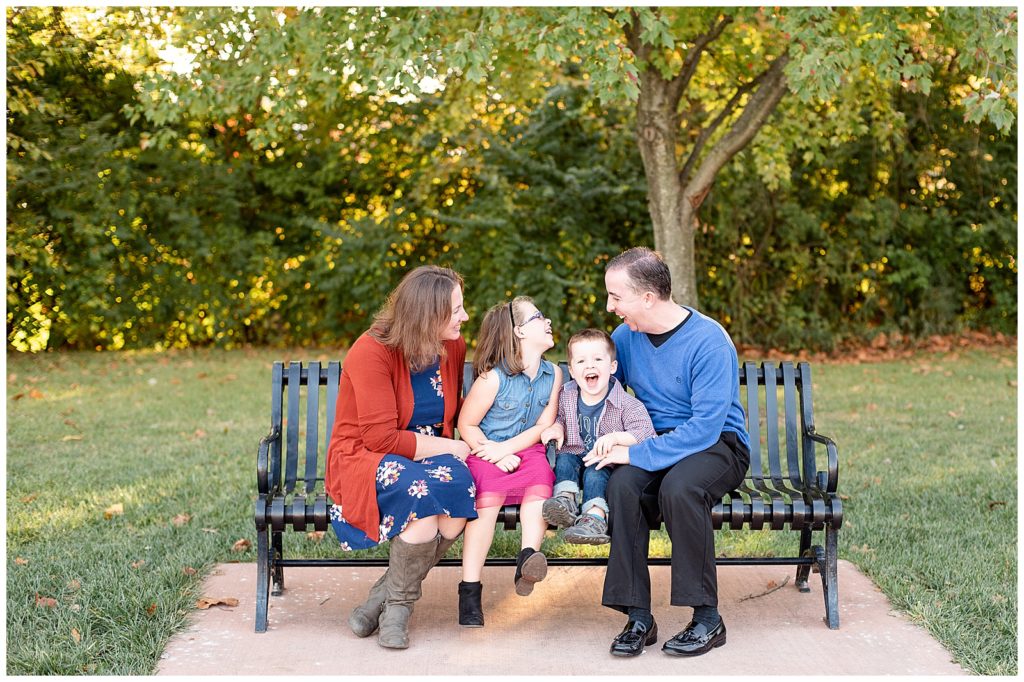 a family of 4 sits on a bench and laughs for fall family photos
